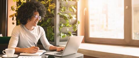 Black millennial guy working in cafe, browsing on laptop and taking notes, empty space photo