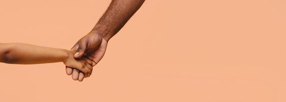 A close-up image of two hands holding each other in front of a peach-colored background. The hands are of different skin tones, representing unity and connection, panorama with copy space photo