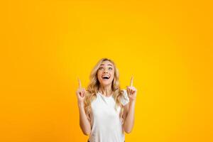 A young woman with long blonde hair is standing against a yellow background. She is pointing upwards with both hands, while smiling and looking up with an excited expression on her face, copy space photo