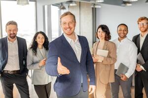 A group of diverse business professionals are standing in a modern office, with one man extending his hand for a handshake. The others are smiling and looking at the camera. photo