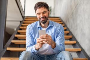 A man in a blue shirt and jeans sits on a wooden staircase in a home, looking down at his smartphone. He appears to be smiling and engrossed in what he is reading or viewing. photo