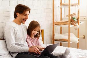Preschool girl spending time with her daddy on weekend at home, using laptop in bedroom, copy space photo