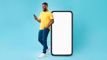 Handsome young Afro man using mobile phone while standing near huge smartphone with empty screen over blue studio background. Mockup for website or application, space for advertisement photo