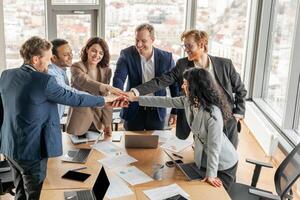 A diverse team of business professionals stand around a conference table, their hands stacked in the center. They smile at each other, showcasing their positive and collaborative work environment. photo