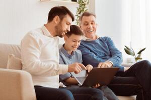 Father And Grandfather Teaching Boy How To Use Laptop And Computer Programs Sitting On Couch At Home. Selective Focus photo