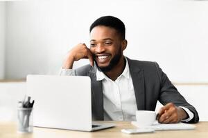A man in a suit smiles as he sits at his desk, looking at a laptop during a call. He has a cup of coffee and a phone on the table in front of him. photo