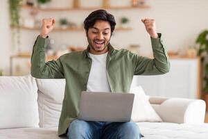 Arab man is seated on a couch, with his arms raised while focused on using a laptop in front of him. He appears engaged and concentrated on the screen, possibly working or browsing the internet. photo