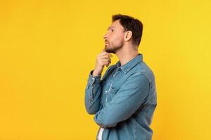 A man standing upright in front of a solid yellow background. The man is facing forward with a neutral expression, touching his chin and looking at copy space photo