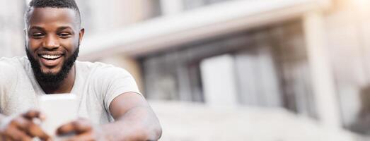 A young man with a beard smiles widely while looking at his phone, sitting on a curb in an urban setting. photo
