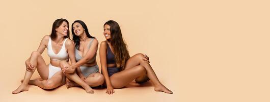 Three glad diverse millennial women in undergarments sit on floor, enjoy body positivity, beauty care, have fun, isolated on light studio background, full length. Friendship, confidence, copy space photo