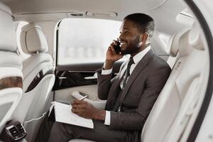Black businessman is sitting in the backseat of a car, taking notes on a notepad while talking on a phone. He is wearing a suit and tie, and he has a serious expression on his face. photo