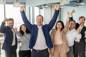 A group of diverse business professionals stand in a modern office, celebrating their success. They are all smiling and laughing, with their arms raised in the air photo