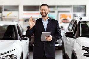 A man standing in front of a car, holding a tablet in his hand. He appears to be inspecting something on the device, with the car parked behind him. photo
