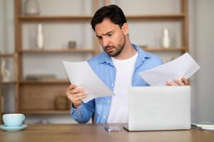 A man is sitting at a desk at home office, looking with a perplexed expression at some documents in his hands. A laptop is open in front of him, suggesting he is cross-referencing information photo