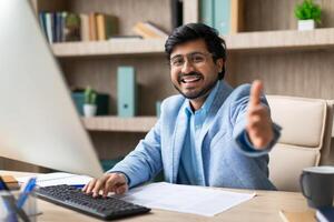 Indian businessman in a blue suit smiles and gives a thumbs up while sitting at his desk and working on his computer. There are papers on his desk, and a bookshelf is visible behind him. photo