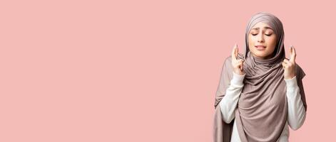 Young muslim girl in hijab crossing fingers, making wish, praying for miracle over pink studio background with free space photo