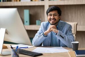 Indian man wearing glasses smiles while sitting at his desk, with a computer and papers in front of him. He has a friendly and confident expression, suggesting he is ready to work or have a meeting. photo
