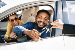 This image depicts a happy couple receiving the keys to their new car from a dealership representative. The man is sitting in the drivers seat of a white sedan and is reaching out to take the keys photo