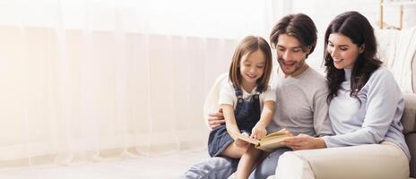 Parents teaching their little daughter how to read, hoding book and smiling while spending time together at home, empty space photo