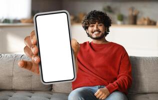 Eastern man is seated on a couch, holding a smart phone in his hand. He appears focused on the devices screen, possibly browsing or messaging. photo