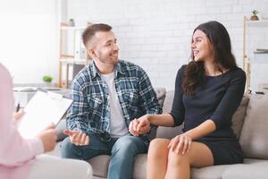 A young couple sits on a couch together holding hands, smiling and looking at each other. They are in a therapy session with a therapist out of frame. The couple appears to be happy and relaxed photo