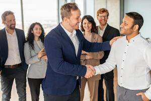 A group of diverse business professionals stand together in a modern office setting. A smiling man in a blue suit extends his hand to shake the hand of a new colleague, who also smiles warmly photo