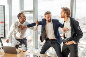 Three men in suits are arguing in a modern office with large windows. One man is pointing and yelling, while another man is gesturing with his hands. The third man is standing back and looking on. photo
