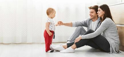 A young family is playing together in their living room. A man and a woman are sitting on the floor, smiling as they watch their baby take his first steps. photo