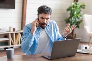 A man appears visibly upset as he argues over the phone, gesturing with his hand. He is seated at a wooden table where his laptop is open, suggesting he is dealing with a work-related issue photo