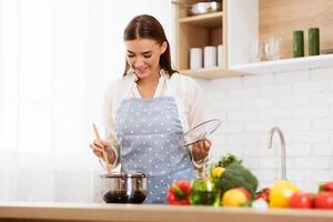 A woman wearing a blue and white apron stirs a pot of food with a wooden spoon, while holding a glass lid in her other hand. Fresh vegetables are on the counter in front of her. photo