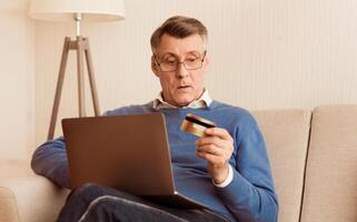 Online Shopping. Senior Man Using Credit Card And Laptop Computer Sitting On Sofa At Home. Selective Focus photo