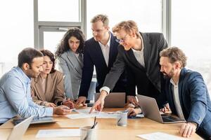 A group of business professionals are gathered around a table in an office setting, discussing a project. The team is reviewing documents and using laptops as they brainstorm ideas and strategize photo