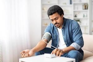 A black man is seated on a couch, focused intently on monitoring his blood pressure using a digital sphygmomanometer. His arm is wrapped in the cuff, connected to a small, portable device photo