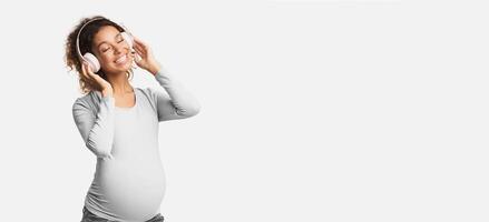 Afro pregnant woman enjoying music in headphones with closed eyes, white studio background photo