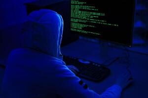 A person wearing a hooded sweatshirt sits in front of a computer with a dark background. Computer screen displays green code, indicating that the person is working on a programming or technical task. photo