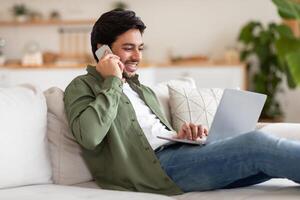 Arab man is seated on a couch, engrossed in his laptop and phone. He appears focused and attentive, balancing both devices on his lap. The room is cozy and well-lit, creating a relaxed atmosphere. photo