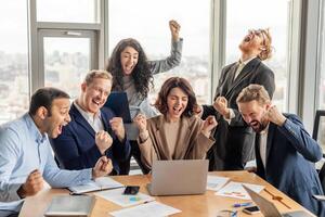 A team of diverse business professionals celebrates success in a modern office setting, with large windows overlooking a city skyline. They are all laughing and cheering, with their fists in the air photo