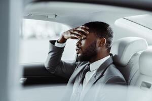 A Black man in a grey suit sits in the back seat of a car. He looks down and has his hand on his forehead as if he is exhausted or stressed. The image is taken from inside the car photo
