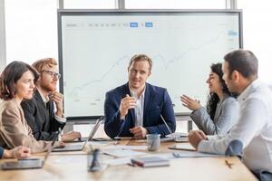 A group of business professionals are seated around a conference table, engaged in a discussion. They are gathered in a modern office with a large whiteboard behind them photo