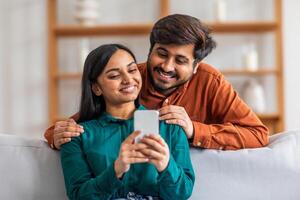 Indian man and woman are sitting together on a couch, both focused on a cell phone in the mans hand. They appear engrossed in the screen, possibly watching something or browsing social media. photo