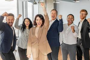 A diverse group of six business professionals, dressed in professional attire, stand together in a modern office setting. They are all smiling and raising their fists in the air photo