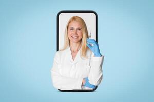 A smiling lady doctor with blonde hair is seen holding a syringe in her right hand while looking at the camera. She is wearing a white lab coat and blue gloves photo