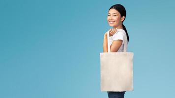 Ecology Concept. Asian Girl With White Eco Bag Smiling At Camera Standing On Blue Studio Background photo