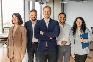 A diverse group of five business professionals stand together in a modern office setting. They are all wearing business attire and appear confident and successful. photo