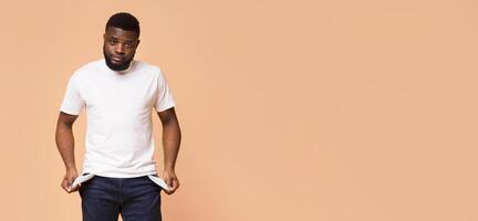 A young black man with a beard is standing against a beige backdrop. He is wearing a white t-shirt and blue jeans and is checking his pockets, which appear to be empty, copy space photo