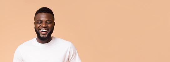 A young African American man with a beard is smiling and looking down. He is wearing a white t-shirt and is standing against a peach background, panorama with copy space photo