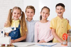 Four young students, two boys and two girls, are smiling and working on a science experiment in a classroom. They are all wearing casual clothing and are standing around a table with a robot photo