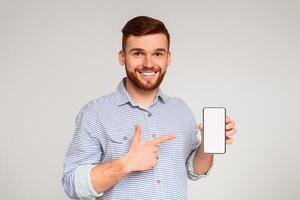 Smiling millennial man holding latest slim cellphone with blank white screen and pointing at it, mockup, free space photo