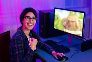 A woman with glasses sits at her computer, smiling and raising her fist in victory while playing a game. The room is lit with purple and blue lights, creating a vibrant and exciting atmosphere. photo