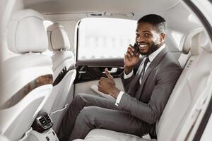 African American successful businessman, dressed in a suit, sits in the backseat of a car. He is talking on his phone, smiling, and giving a thumbs up. The car is clean and luxurious photo